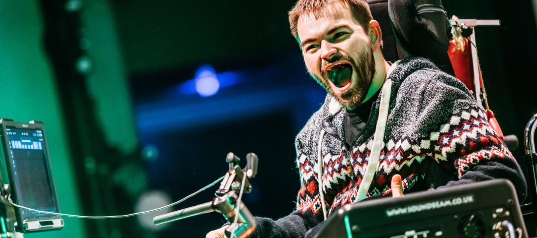 Male adult with beard sat in wheelchair with mouth wide open performing on stage