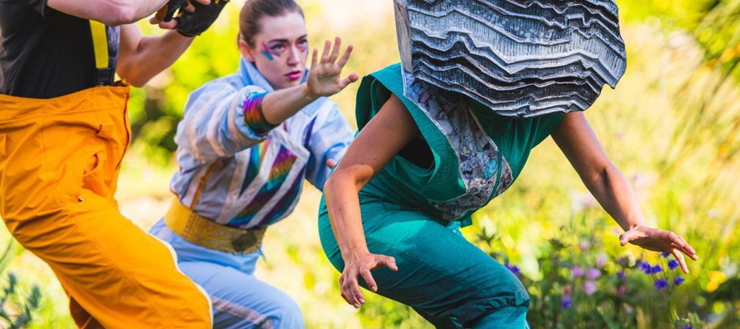 A group of 3 performers wearing bright jumpsuits with one wearing a large grey rock over their head