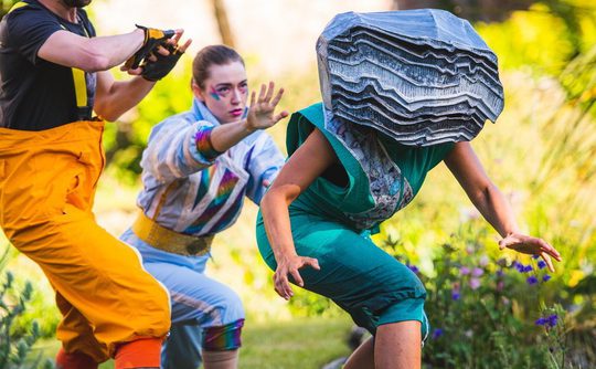 A group of 3 performers wearing bright jumpsuits with one wearing a large grey rock over their head