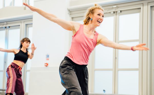 Dance teacher in studio wearing pink top with blonde hair with arms out smiling