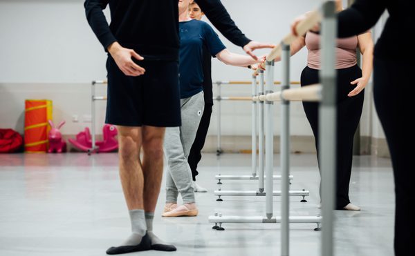 Group of adults standing at a ballet barre in a studio, practicing basic ballet positions with focused posture.