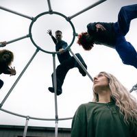 A performer in green looking up at 3 dancers in black climbing on a cage above them