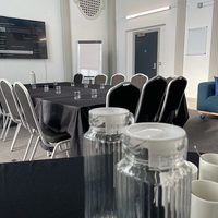 Black meeting tables, black chairs, water jugs, white mugs and a blue couch in the background