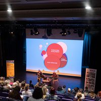 Audience facing two people presenting on stage with projected backdrop of a chart