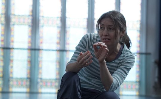 Holly is pictured kneeling in front of a large stained glass window wearing a green and white striped sweatshirt. Her hands are held in front of her in an expressive gesture, thumb and forefinger of one hand touching as she describes something fine or delicate.