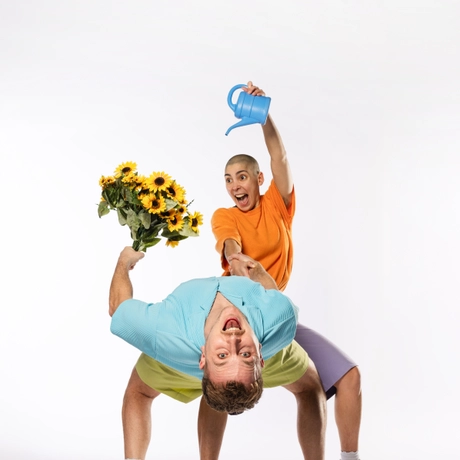 two performers one wearing a blue top and holding a bunch of flowers bending over backwards and one performer wearing an orange top pouring out of a watering can over the flowers