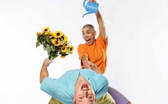 two performers one wearing a blue top and holding a bunch of flowers bending over backwards and one performer wearing an orange top pouring out of a watering can over the flowers
