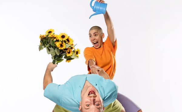 two performers one wearing a blue top and holding a bunch of flowers bending over backwards and one performer wearing an orange top pouring out of a watering can over the flowers