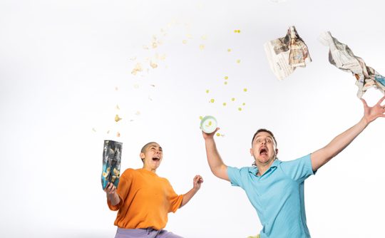 two performers sat on the floor one in an orange top and one in a blue top throwing things in the air